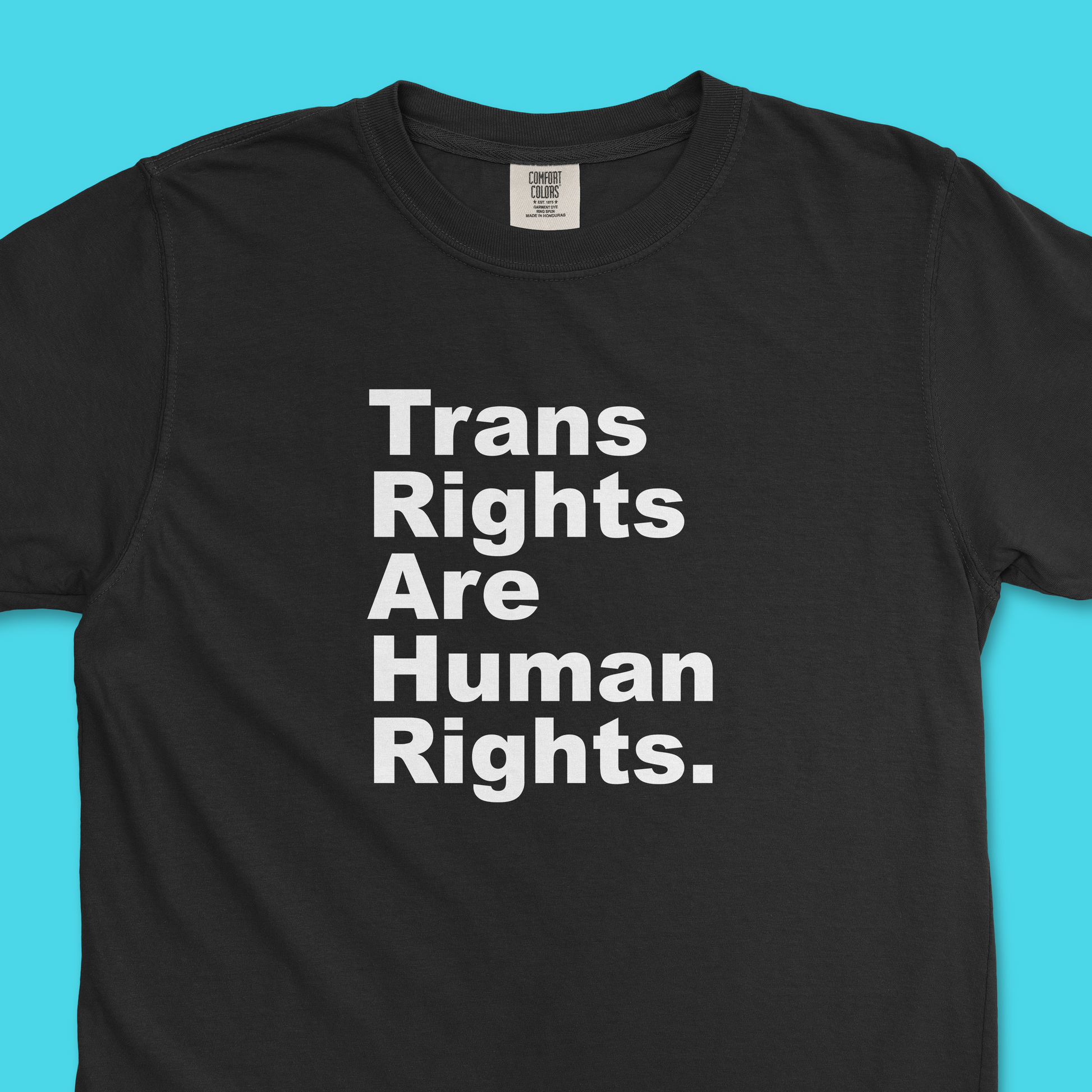 Closeup of black Comfort Colors tee with large white text that says "Trans Rights Are Human Rights" with each word on a separate line and left aligned. Background is light blue.